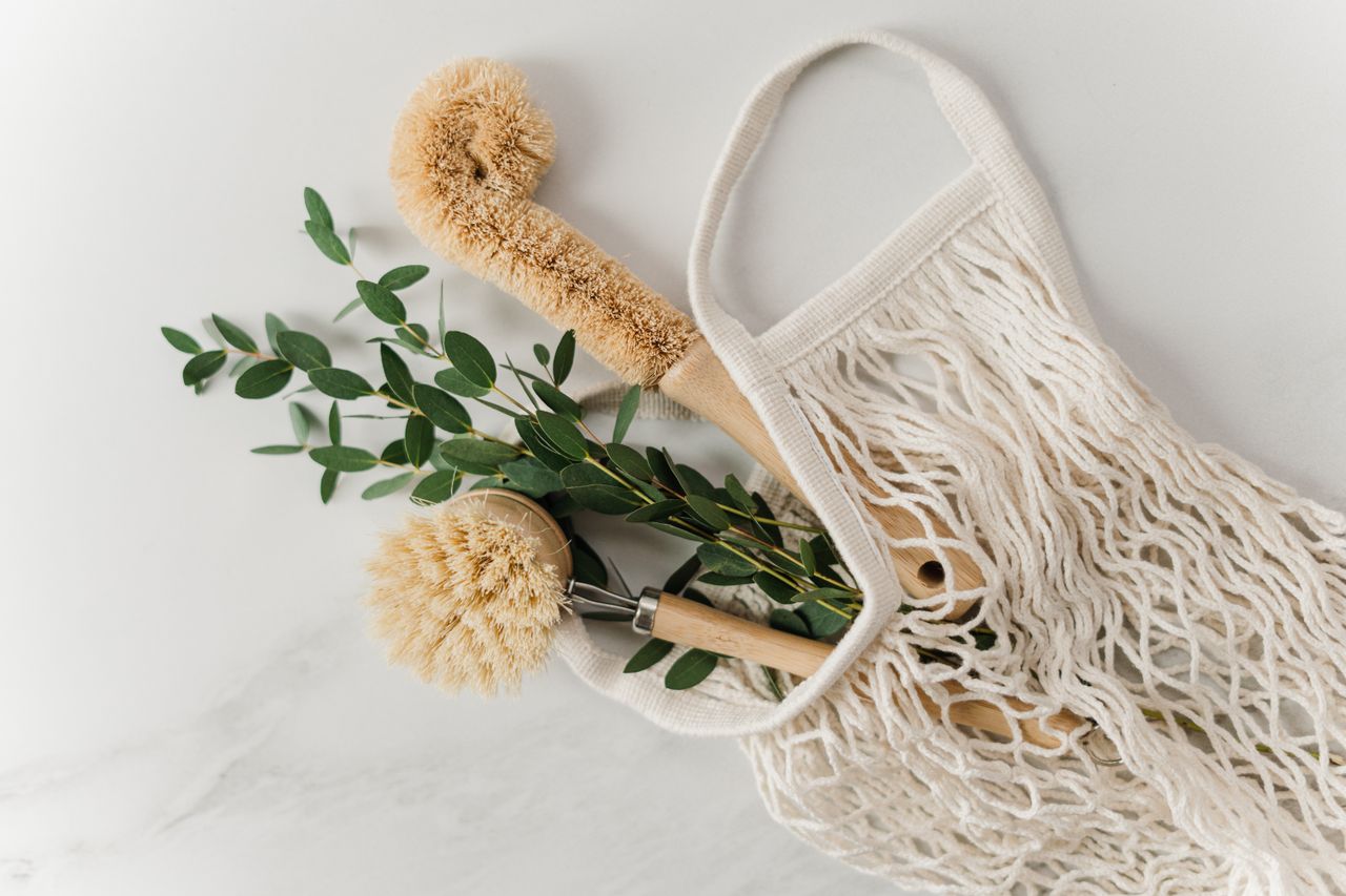 Eco-friendly cleaning brushes and eucalyptus branch in a net bag on a marble surface.